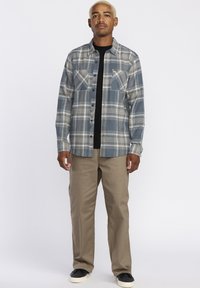 Chemise en flanelle à carreaux gris avec deux poches poitrine, t-shirt noir à col rond en dessous, pantalons beiges et baskets noires. Texture lisse et fermeture à boutons.