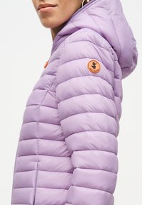 Άτομο που φοράει ένα ανοιχτό μωβ κοντό μπουφάν με κουκούλα και πορτοκαλί στρογγυλή ραφή στο μανίκι με λογότυπο πάπιας.