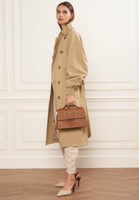 Trench coat beige con grandi bottoni, abbinato a pantaloni tapered chiari e décolleté beige con tacco. Borsa strutturata marrone con chiusura.