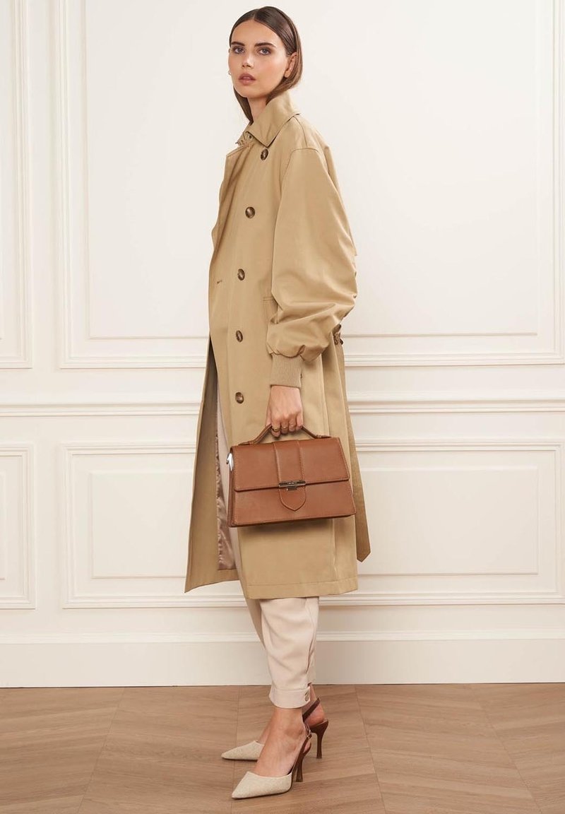 Trench coat beige con grandi bottoni, abbinato a pantaloni tapered chiari e décolleté beige con tacco. Borsa strutturata marrone con chiusura.