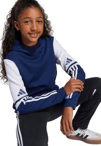Haut de survêtement bleu marine et blanc avec un col montant, présentant trois rayures blanches sur les manches. Pantalon noir avec des rayures blanches sur les côtés et des chaussures grises.