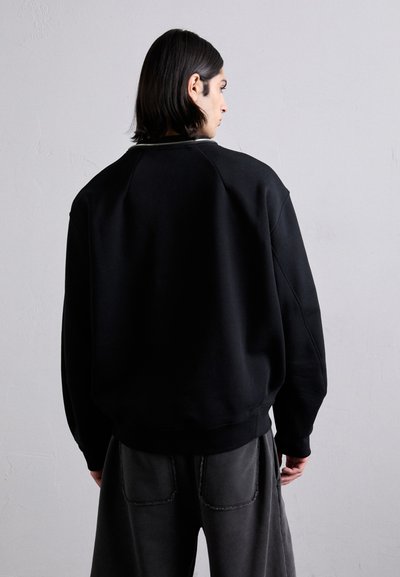 Črna športna majica z raglan rokavi, izdelana iz gostega bombažnega materiala. Ima rebrast ovratnik in manšete, v kombinaciji s prostornimi, sivimi hlačami.