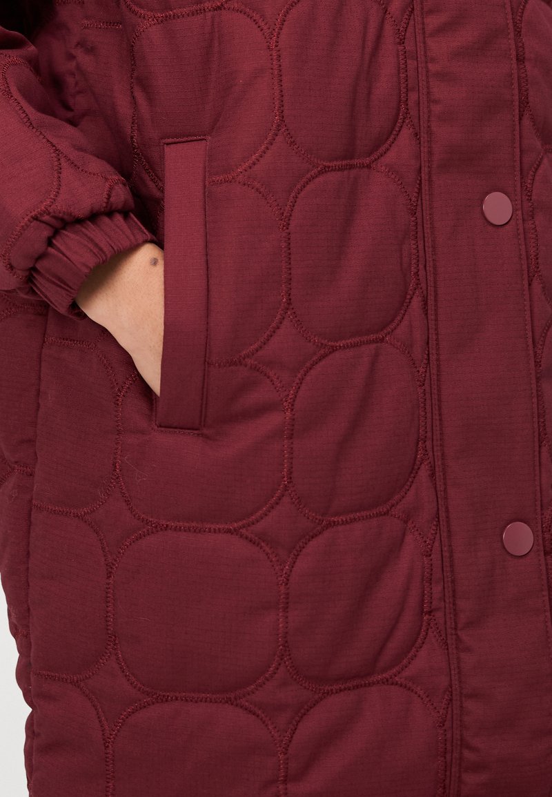 Veste matelassée bordeaux avec des motifs de couture circulaires, dotée d'une poche latérale et de poignets élastiques pour un ajustement confortable.