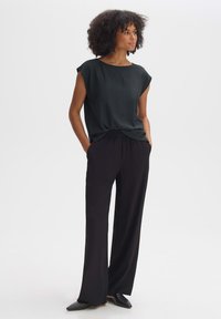 Top strutturato verde scuro con maniche corte infilato in pantaloni neri a vita alta e larghi; modella che indossa scarpe nere slip-on.