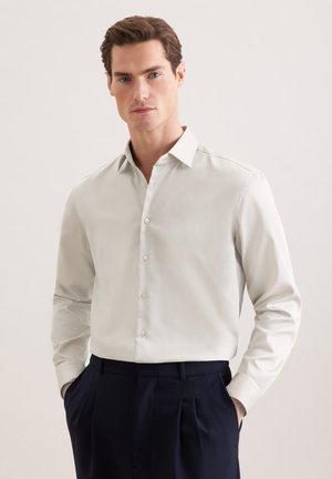 Homme portant une chemise blanche boutonnée à manches longues et un pantalon sombre, debout les mains dans les poches devant un fond clair et uni.