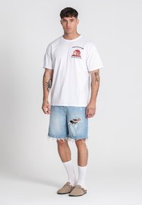 Camiseta blanca de algodón con un estampado gráfico y pantalones cortos de mezclilla azul con bordes deshilachados y desgaste; complementados con zapatillas beige sin cordones.