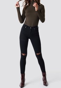 Czarne dżinsy z wysokim stanem i przetarciami na kolanach, zestawione z dopasowanym oliwkowym golfem. Model ma na sobie buraczkowe botki za kostkę. Gładka faktura.