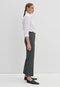 Chemise blanche à boutonnière associée à un pantalon taille haute gris sur mesure et des chaussures plates noires. La chemise a des manches retroussées et une coupe décontractée.