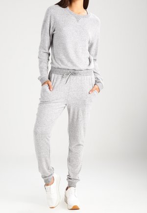 Frau trägt ein hellgraues Sweatshirt mit langen Ärmeln und passende Jogginghose sowie weiße Turnschuhe, steht mit den Händen in den Taschen.