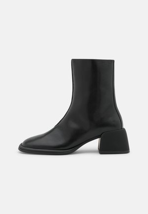 Bota de tornozelo em couro preto com biqueira pontiaguda e um salto geométrico robusto. Textura suave com detalhes mínimos.