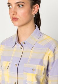 Lilla og gul rutete skjorte med krage, to brystlommer og detaljer med sølvknapper. Stoffet har en teksturert overflate.