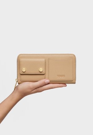 Mano sosteniendo una cartera de cuero beige con bolsillo frontal con cierre de botón a presión, ranura para tarjetas, superficie texturizada y logo de TOUS en relieve sobre fondo blanco.