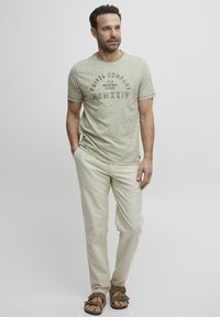 Šviesiai žalia melange t-shirt su apvaliu kaklu, juodu piešiniu, smėlio spalvos kelnėmis ir rudomis šlepetėmis. Atsitiktinis, laisvas kirpimas.