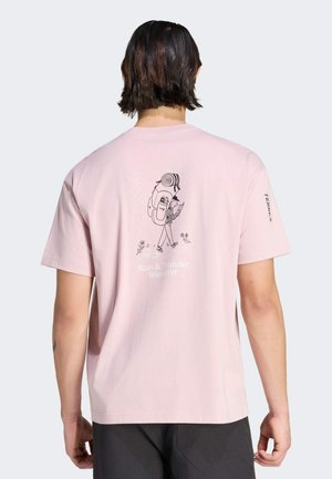 Persona con capelli scuri fino alle spalle che indossa una T-shirt rosa chiaro con grafica da escursionismo e la scritta "Rain & Thunder Wanderer" sulla schiena.