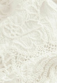 Tissu blanc avec des motifs de dentelle en relief complexes et des boucles créant un design floral et tourbillonnant texturé.