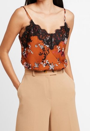 Vrouw draagt een mouwloos oranje bloemenkamisol met zwarte kantafwerking, ingestopt in beige wijde broek met hoge taille, handen in de zakken.