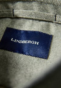 Marine stof etiket genaaid op lichtgrijze denim, met het woord "LINDBERGH" in witte hoofdletters, en zichtbare stikseldetails.