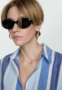 Ochelari de soare ovali negri, cămașă cu dungi albastre și albe, cercei rotunzi din aur, texturi netede, design casual, cu părul scurt blond.