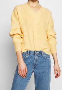 Pull en maille torsadée avec col en V, de couleur jaune doux, manches amples et ourlet côtelé. Assorti à un jean en denim bleu clair.