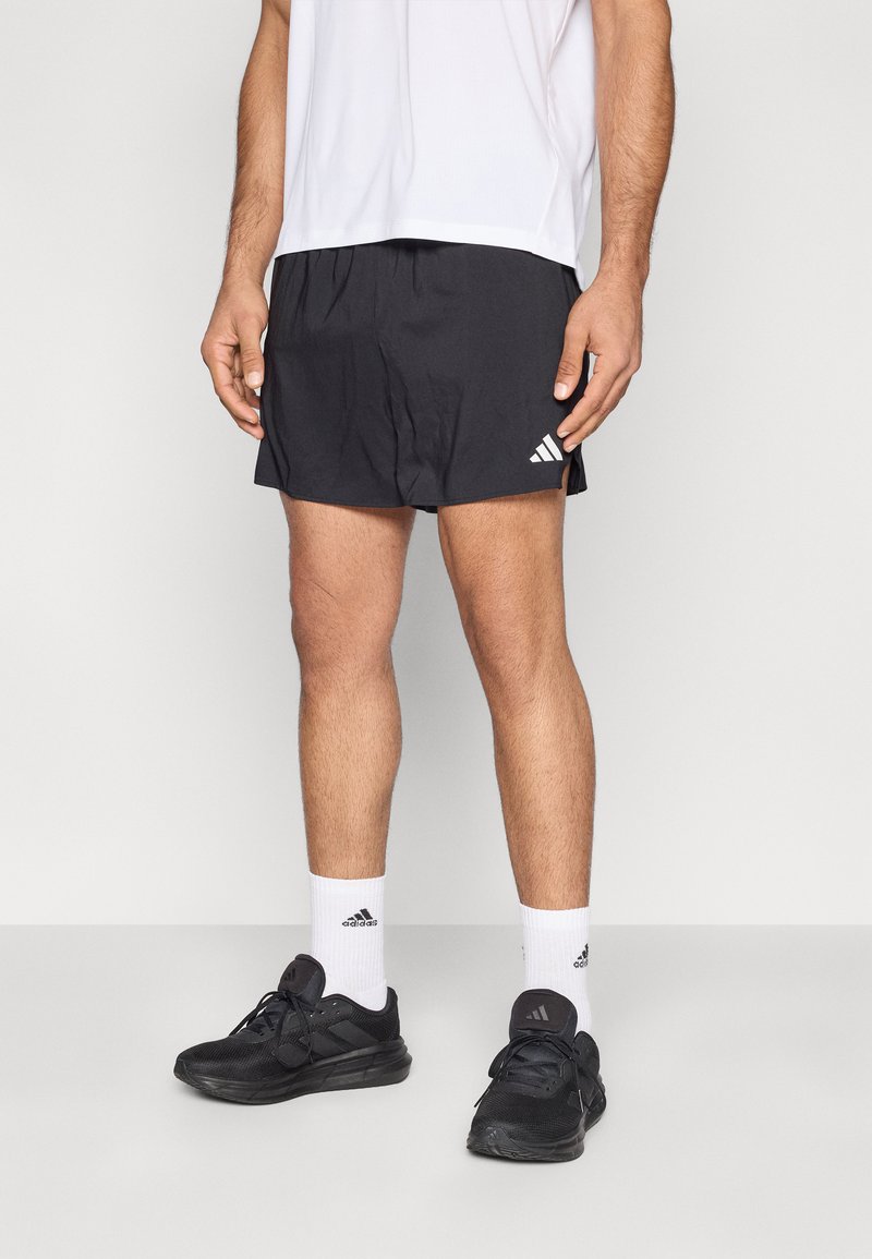 Shorts de sport noirs avec taille élastique et fentes latérales, associés à un t-shirt blanc et des chaussures de course noires. Chaussettes blanches avec logo visible.