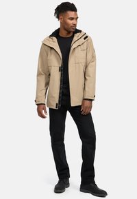 Beige waterdichte jas met een zwarte binnenkant en een verstelbare capuchon. Voorzien van voorzakken en een ritssluiting met zwarte accenten.