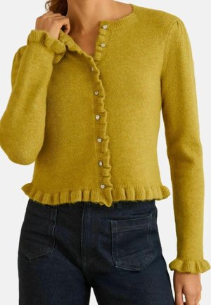Femme portant un cardigan jaune moutarde avec des bords volantés et des boutons en forme de cœur, associé à un jean foncé taille haute.