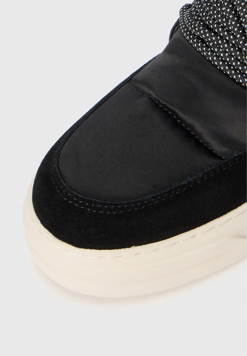 Zapatilla negra con parte superior de satén y acentos de ante, con un detalle de cordones texturizados en gris y una suela de goma color crema.