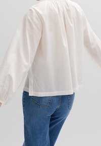 Personne portant une blouse blanche ample à manches longues avec une texture subtile sur les manches et un jean en denim bleu, vue de dos.