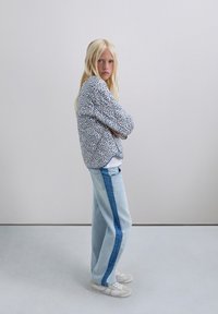 Chaqueta con estampado floral y acentos en azul y rojo, combinada con jeans azul claro que presentan una franja lateral azul oscura, y zapatillas blancas.