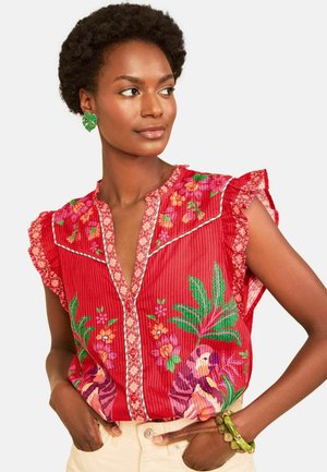 Woman with short curly hair wearing a red floral patterned blouse, green leaf earrings, beige pants, and green bracelets, looking sideways.