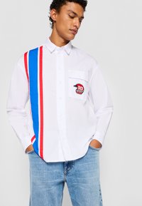 Hvid skjorte med knapper, der har blå og røde lodrette striber, en brystlomme med et sort og rødt logo-patch, og afslappede denim jeans.