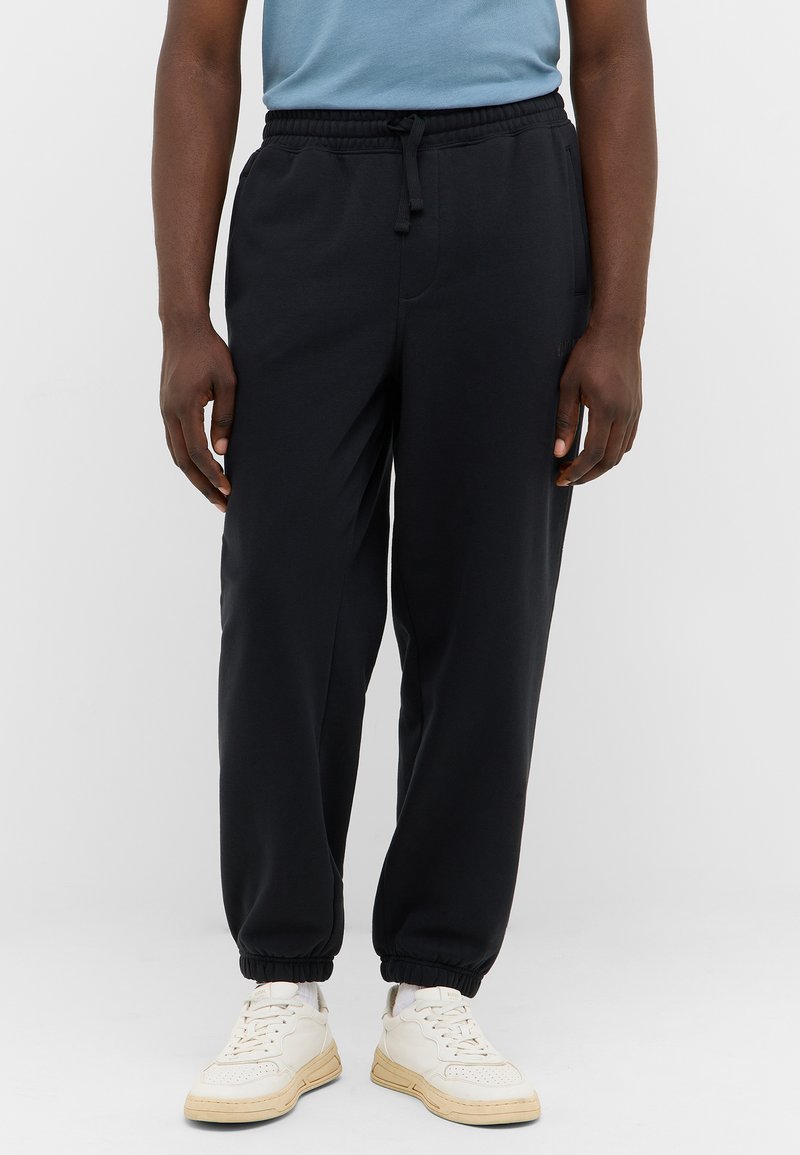 Schwarze Sweatpants mit elastischem Bund und Bündchen, die einen lässigen Schnitt und eine weiche Textur aufweisen. Tragen mit weißen Sneakers.