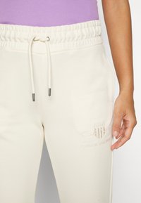 Pantaloni sportivi color crema con vita elastica e cordino. Presentano un logo ricamato sulla coscia e tasche laterali. Tessuto morbido.