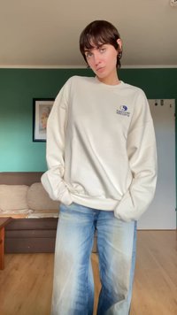 Sweatshirt de cor creme com um corte largo, apresentando um logótipo azul no lado esquerdo, combinado com jeans de perna larga azul desbotados.