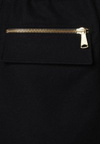 Zwarte stof met een gladde textuur en een horizontale zak, aangevuld met een gouden ritssluiting die een contrasterend metallic detail toevoegt.