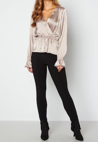 Blusa avvolgente in satin color taupe con maniche lunghe, scollatura a V, vita elastica e polsini arricciati, abbinata a pantaloni neri aderenti e tacchi.