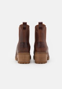 Καφέ δερμάτινα μποτίνια μέχρι τον αστράγαλο με υφή, χ�αρακτηριστικά μπλοκ τακουνιού, θηλιές για εύκολη χρήση και ένα λογότυπο στο πίσω μέρος.