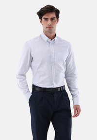 Weiß-blau gestreiftes Hemd zum Knöpfen, lange Ärmel, eine Brusttasche, kombiniert mit dunkelblauen Hosen und einem schwarzen Gürtel.