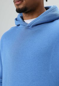 Blauwe hoodie van zachte, gestructureerde stof, met een verstelbare capuchon en een eenvoudig ontwerp. De kleur is egaal, zonder zichtbare patronen.