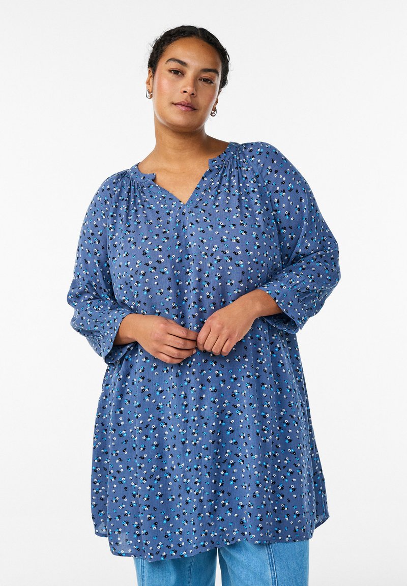 Frau trägt eine locker sitzende blaue Bluse mit Blumenmuster, 3/4 Puffärmeln und hellblauen Jeans, steht mit vor dem Körper verschränkten Händen.