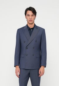 Giacca in tessuto testurizzato blu navy a due bottoni con rever a punta, bottoni neri e due tasche frontali, abbinata a una camicia scura.