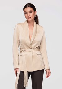 Femme portant un blazer beige ceinturé et un pantalon noir, posant devant un fond gris clair, avec des boucles d'oreilles créoles dorées et un maquillage naturel.