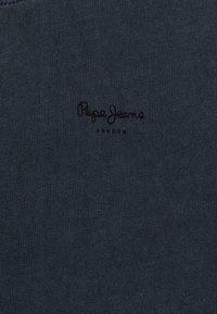 Tmavě modrá bavlněná tkanina s hladkým povrchem, na jejímž vrchu je černě vyšitá logo "Pepe Jeans".