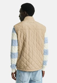 Beige gesteppte Weste mit ärmellosem Design, featuring strukturierte blau-weiße gestreifte Ärmel aus Zopfmuster und gerippte Bündchen.
