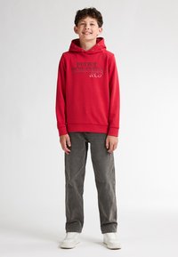 Felpa rossa con logo stampato grigio, dotata di cappuccio con cordino. Abbinata a jeans in denim grigio e sneakers bianche.