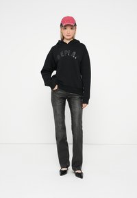 Schwarzer Hoodie mit "REPLAY"-Logo, schwarze Jeans und einer rosa Kappe. Die Hose hat eine ausgewaschene Textur, und die Schuhe sind schwarz mit Schleifen.