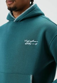 Een tealkleurige hoodie gemaakt van zachte stof, met witte decoratieve stiksels rond de capuchon en een geborduurd logo op de borst.