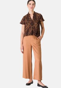 Blouse met luipaardprint, korte mouwen, zwart en bruin, knoopsluiting aan de voorkant. Brede tan-kleurige broek, gladde textuur, normale taille met zijzakken, platte zwarte schoenen.