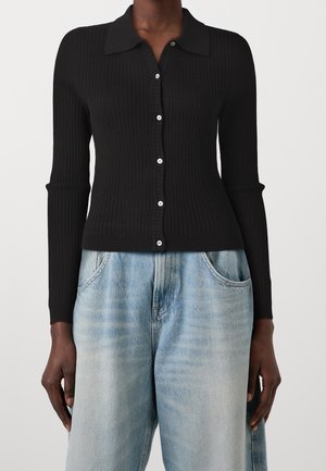 Zwarte geribbelde cardigan met knopen en lange mouwen, gecombineerd met lichtblauwe, hoog gesloten loszittende jeans gedragen door een staand persoon.