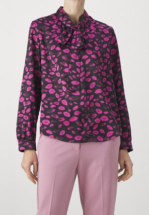Blouse noire avec un imprimé de lèvres roses, présentant des manches longues, un détail de nœud au cou et une texture lisse et soyeuse. Associée à un pantalon rose.